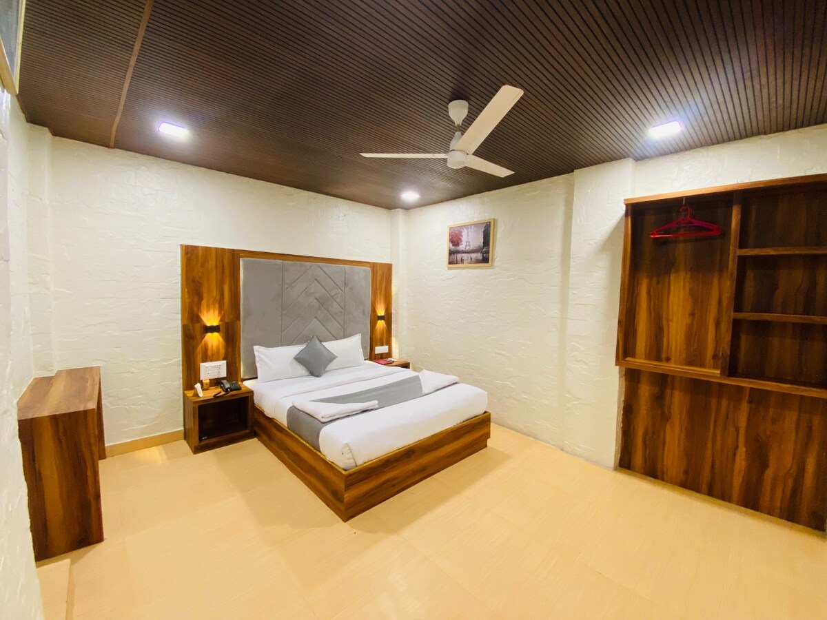 The room features a spacious bed with a light-colored blanket, situated against a wood-paneled headboard. Soft lighting is provided by mounted wall sconces on either side. A wooden dresser and a shelving unit are visible, with a small wall-mounted artwork enhancing the decor.