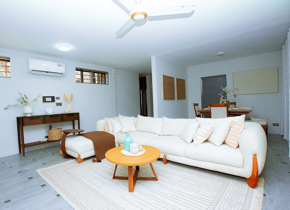 A light-filled living area features a spacious white sofa with wooden legs, complemented by a circular wooden coffee table. An air conditioning unit is mounted on the wall, and a dining area is visible in the background, furnished with a table and chairs.