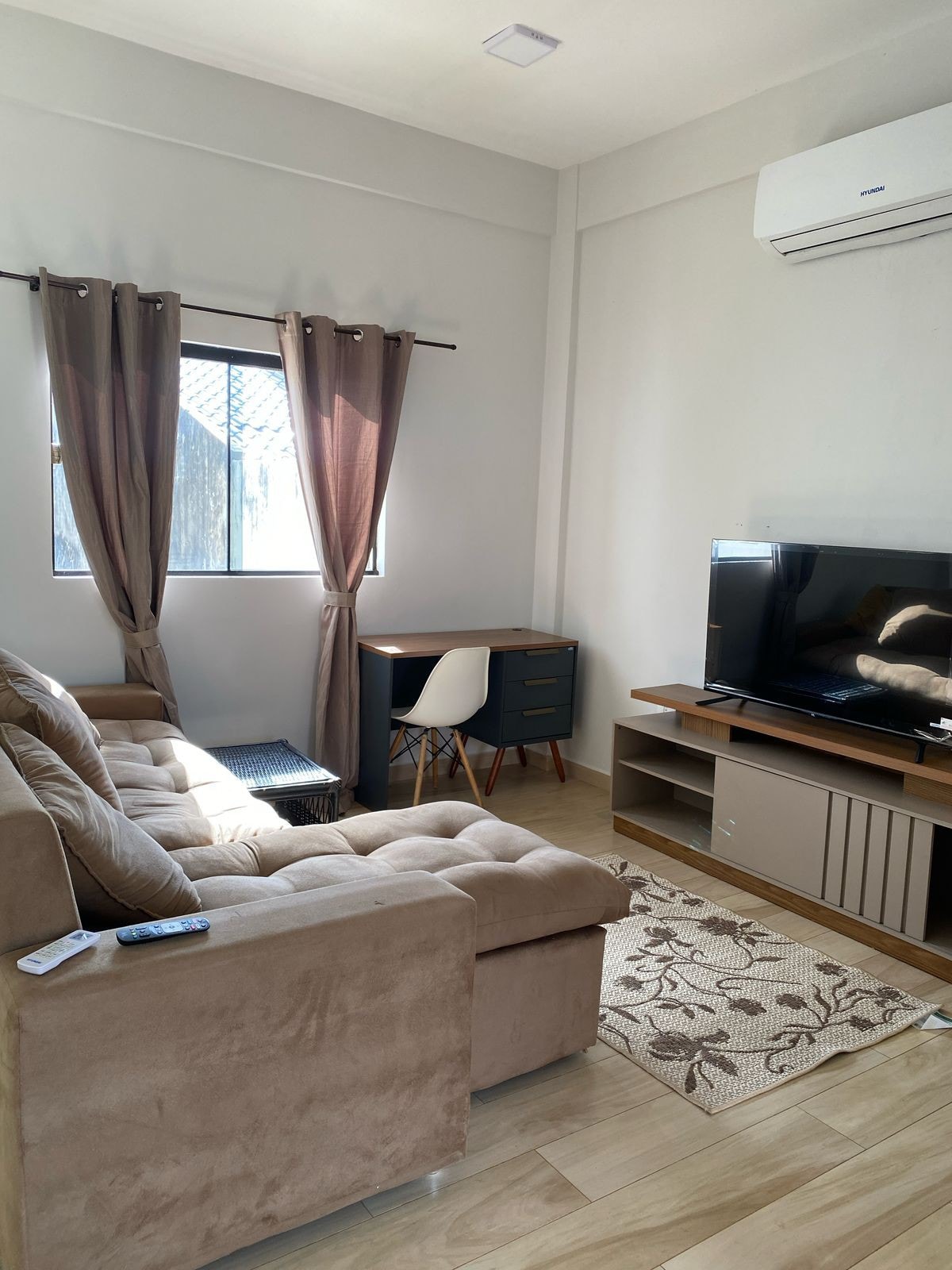 A cozy living area features a plush sectional sofa and a sleek television stand. Natural light enters through large windows adorned with curtains. A small desk is positioned nearby, contributing to a functional workspace. The warm wood flooring creates a welcoming atmosphere.