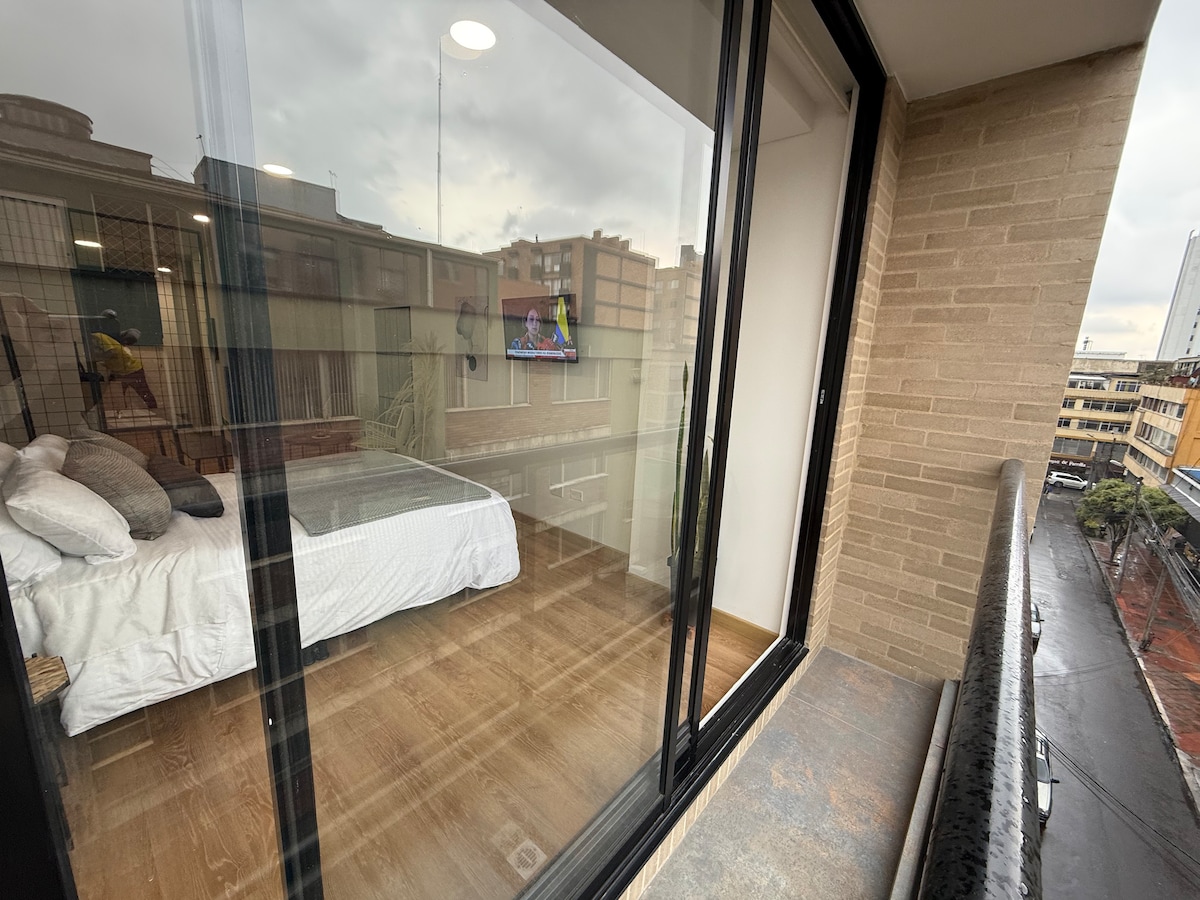 A balcony with a spacious view features glass doors that open to a well-lit room. Inside, a large bed is visible, complemented by soft bedding. The wooden floor contrasts with the light-colored walls, bringing warmth to the space. The scene reflects elements of the city beyond.