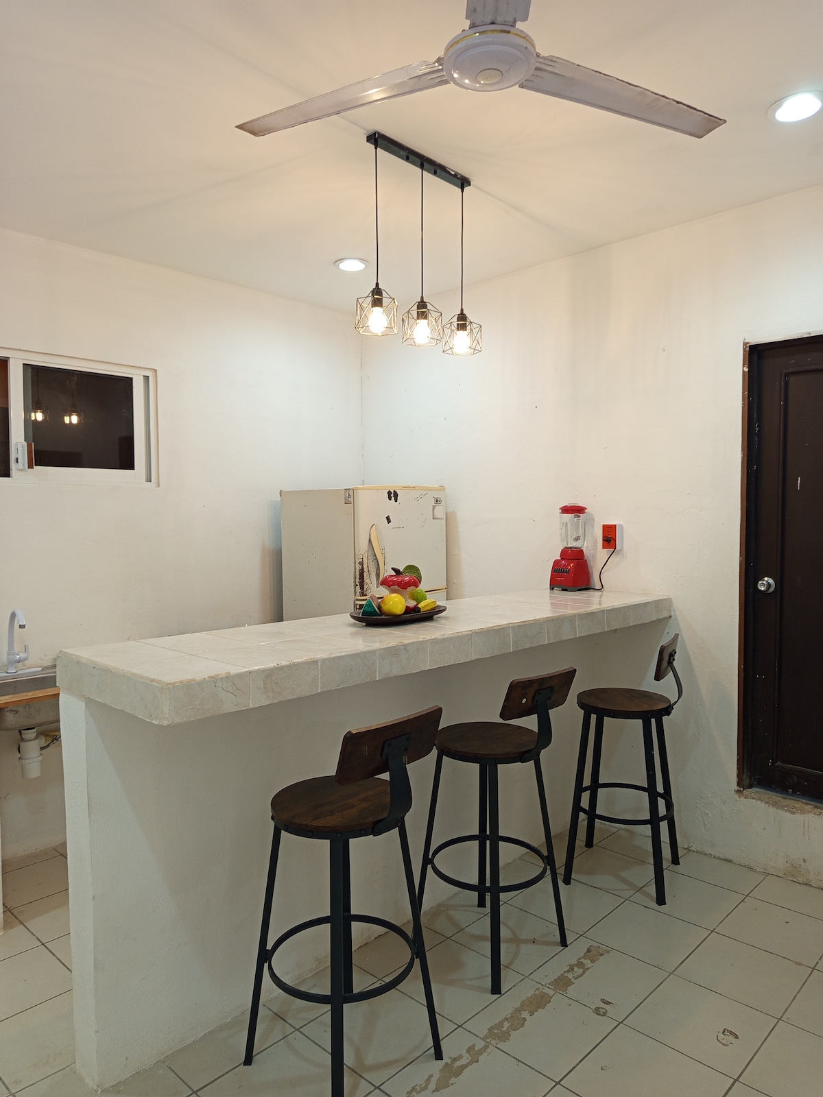 A functional kitchen space features a long counter with three wooden stools, illuminated by pendant lights. A blender is positioned on the counter, alongside a bowl of fruit. A small window allows natural light to enter, and a door is visible on the right.