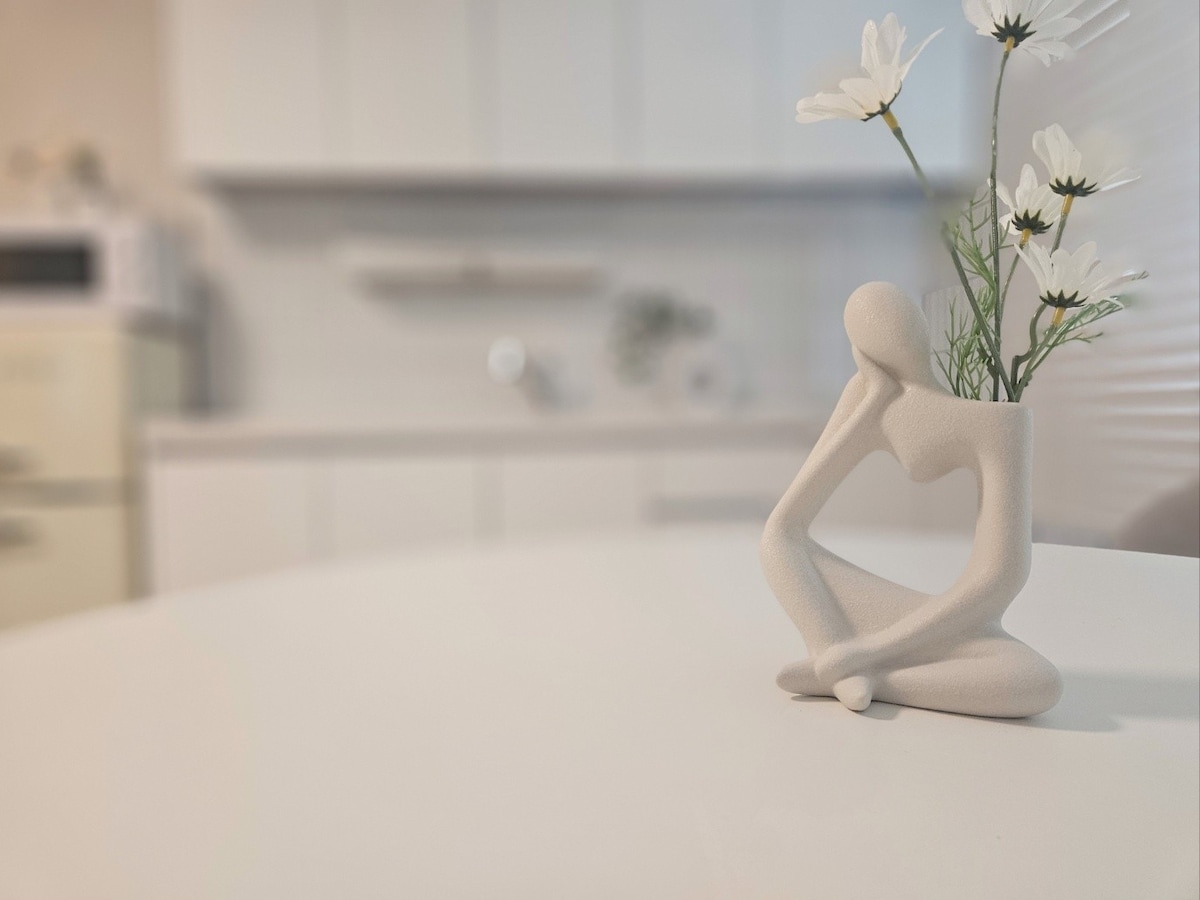 A minimalist figurine, depicting a seated figure, is positioned on a round table. It holds a small arrangement of white flowers, adding a touch of nature to the clean, modern kitchen backdrop with soft colors and minimalist décor.