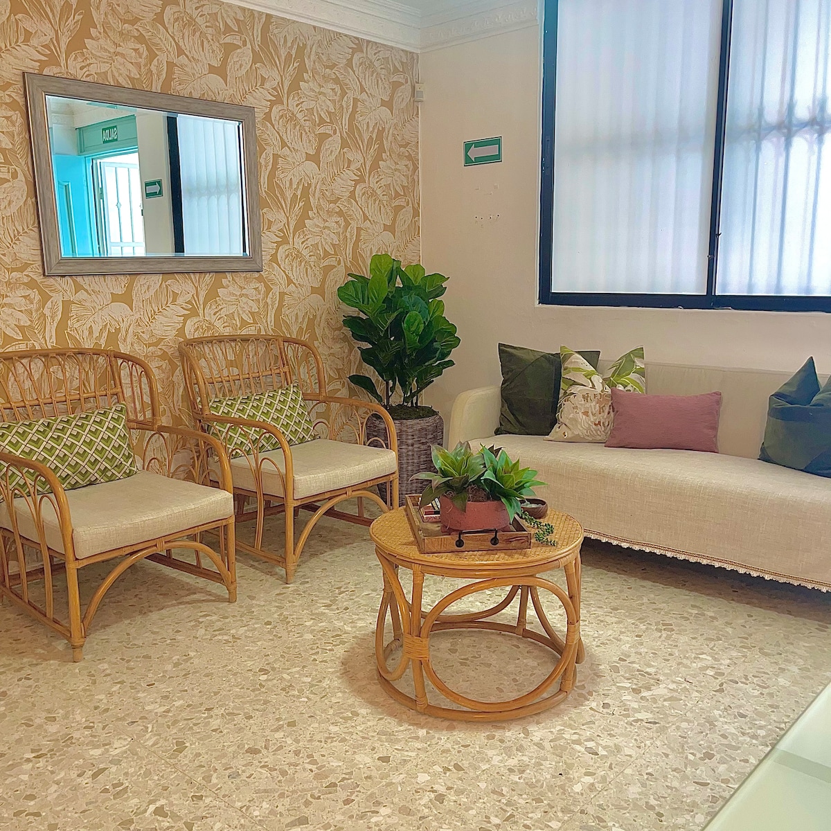 A welcoming reception area features rattan seating with patterned cushions. A round coffee table with a small plant arrangement is centered in the space. Natural light is filtered through a large window, complementing the soft tones of the wall adorned with a floral design.