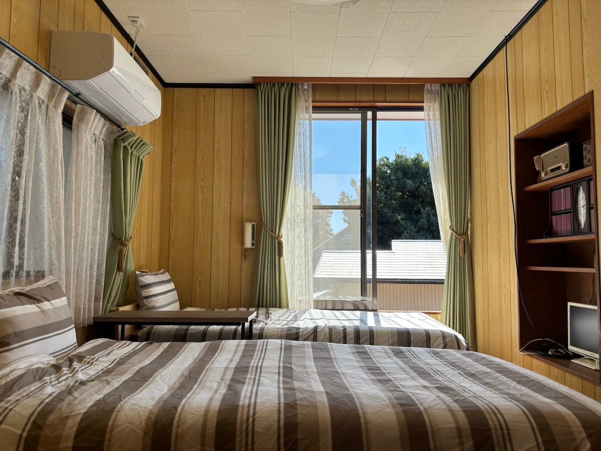 A bright room features two beds dressed in striped bedding, with large windows allowing natural light to fill the space. Green curtains frame the windows, and a small desk is placed against the wall. A bookshelf and a television are present in the corner.