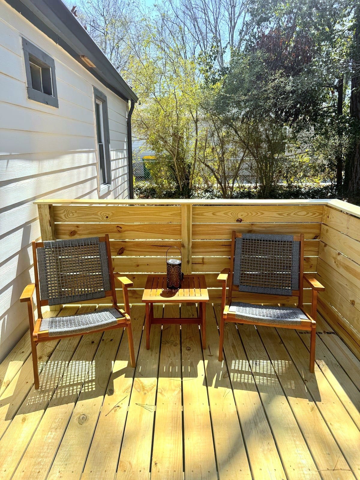 An inviting outdoor deck is showcased, featuring two wooden chairs with woven seats and a small table positioned between them. Natural light filters through nearby trees, enhancing the peaceful atmosphere of the space.