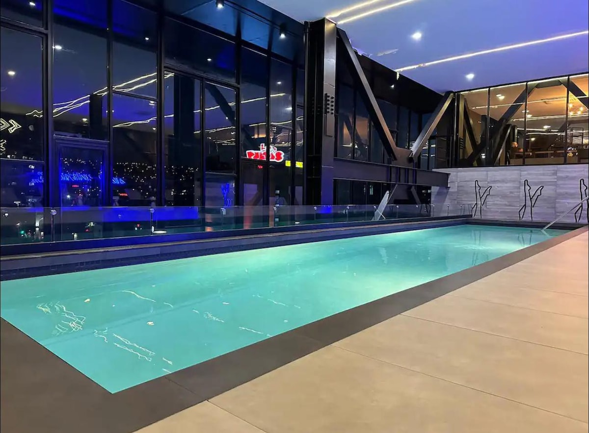 A modern indoor pool is depicted, featuring clear turquoise water under ambient lighting. Expansive glass walls provide views of the surrounding area, while the sleek architectural design includes a minimalist aesthetic with polished surfaces. The atmosphere is tranquil, ideal for relaxation.