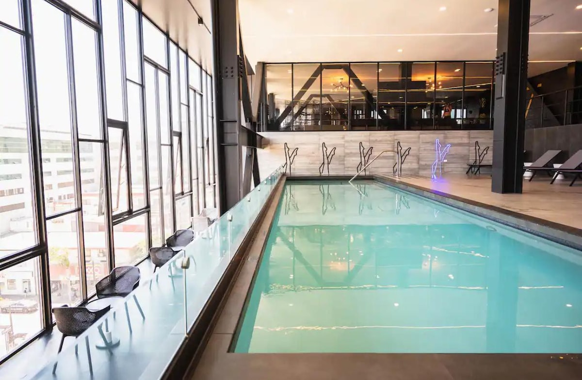A modern indoor pool area features floor-to-ceiling windows that provide ample natural light. The water appears clear and inviting, complemented by sleek, minimalist decor. Several lounge chairs are positioned along the edge, creating a relaxed atmosphere for guests to enjoy.