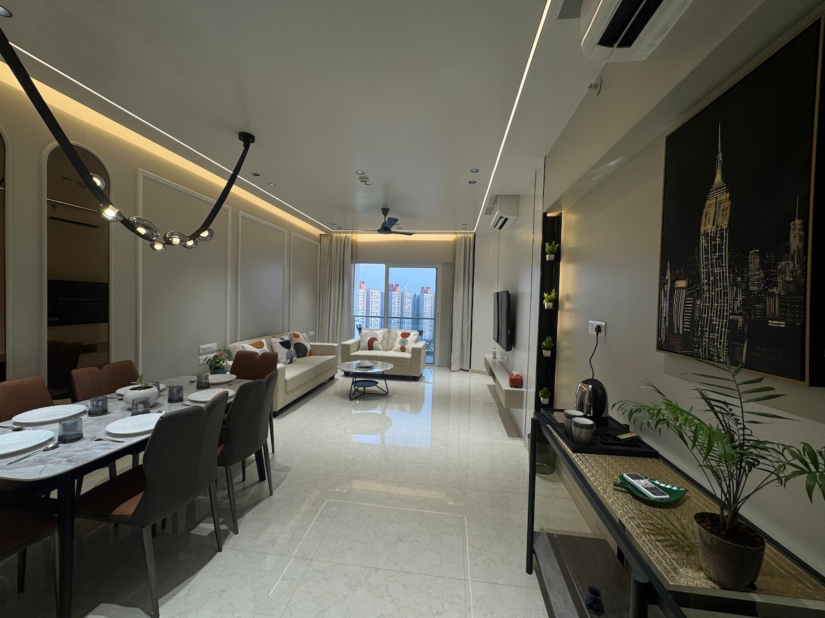 A spacious living and dining area features a sleek design with modern furnishings. A large dining table is set for six, while a comfortable seating arrangement is positioned for relaxation. Natural light enters through wide glass doors, offering views of the skyline.