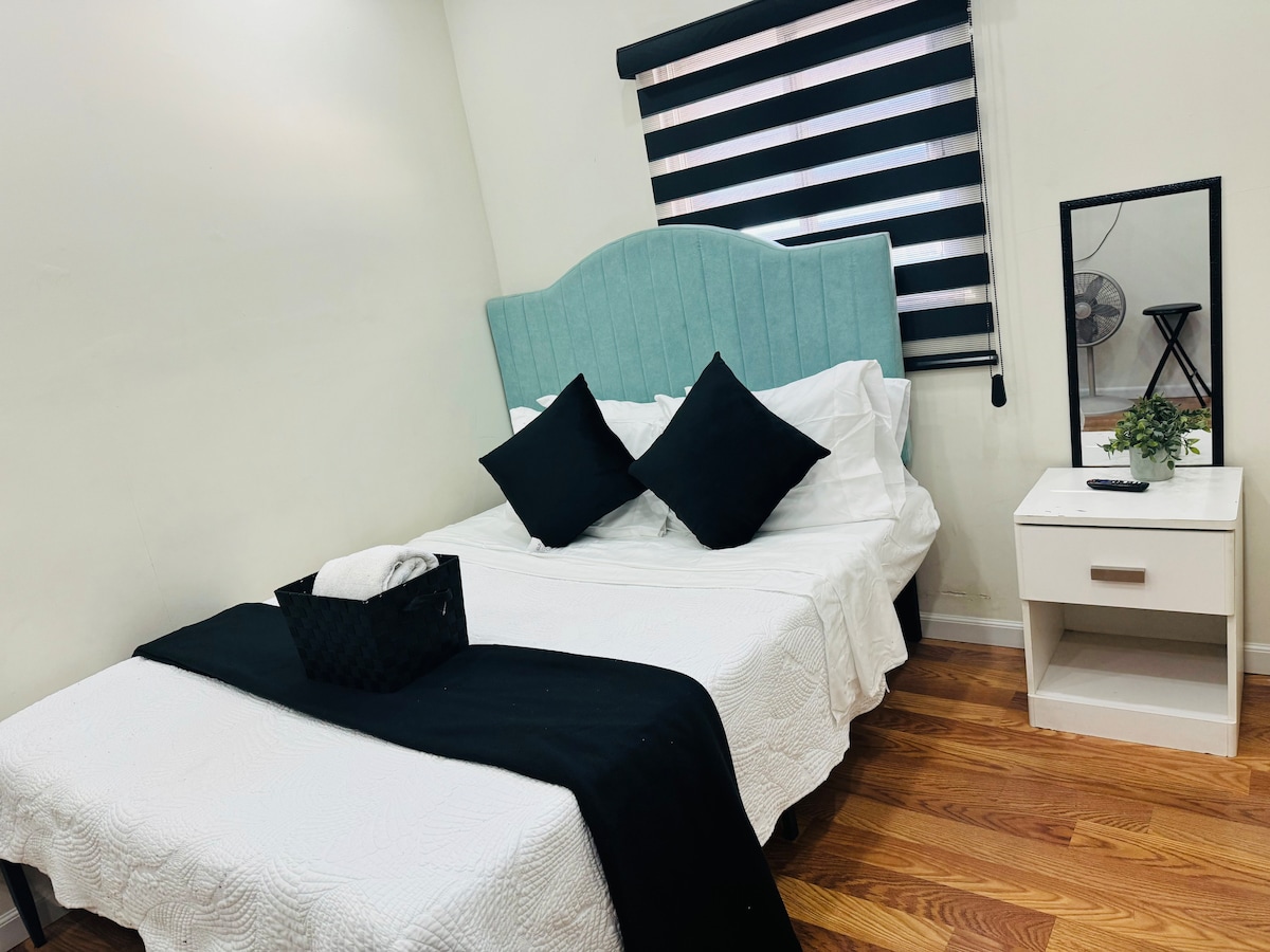 A clean and spacious sleeping area is equipped with a light blue headboard. A white bedspread covers the bed, complemented by black accent pillows. A minimalist nightstand holds a small plant, and a framed mirror reflects natural light from a nearby window with black and white striped blinds.