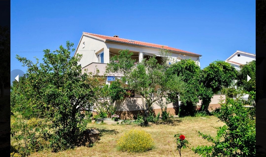 A two-story house is set among lush greenery and blooming flowers, with a spacious balcony overlooking the yard. The surrounding trees and plants contribute to a serene outdoor environment, providing ample space for relaxation.
