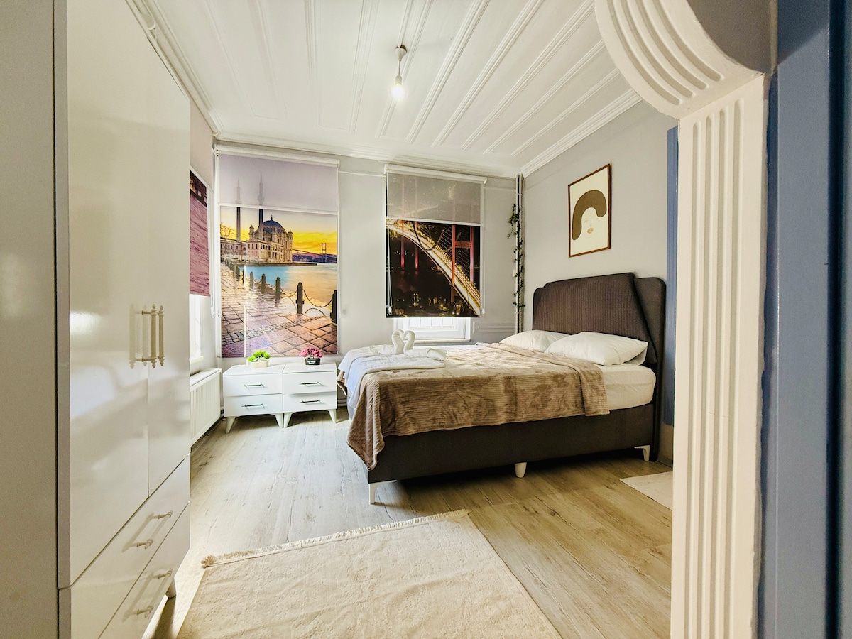 A spacious bedroom features a large bed covered with a neutral-toned blanket. A scenic wall art piece depicting a waterfront view adds interest. Natural light enters through two window coverings, while modern storage options are visible along the opposite wall.