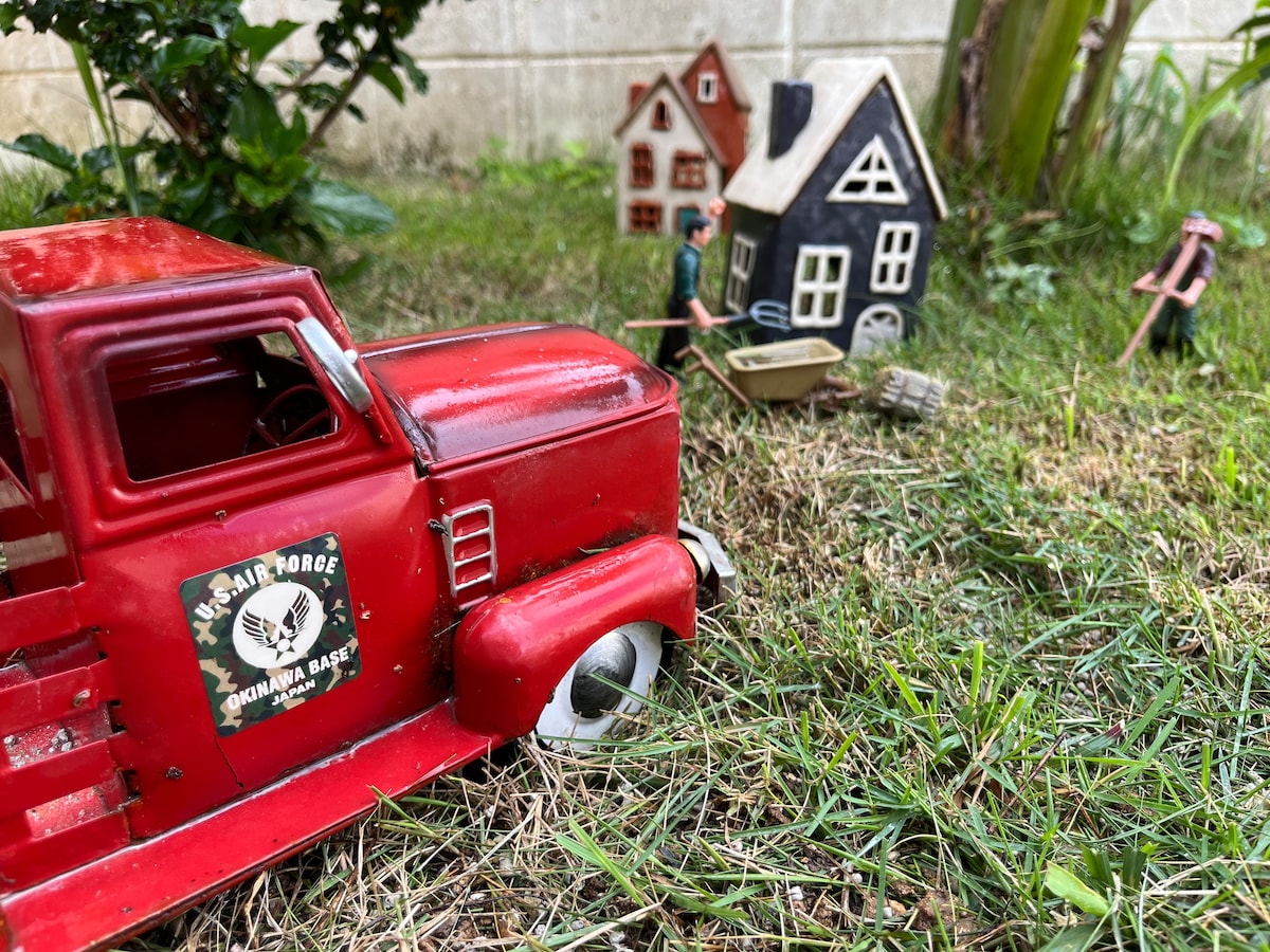 A vintage red toy truck sits in a grassy area, with miniature houses in the background. One house is black with white trim, while the other features a red roof. A small figure and a wheelbarrow are positioned nearby, creating a playful scene.