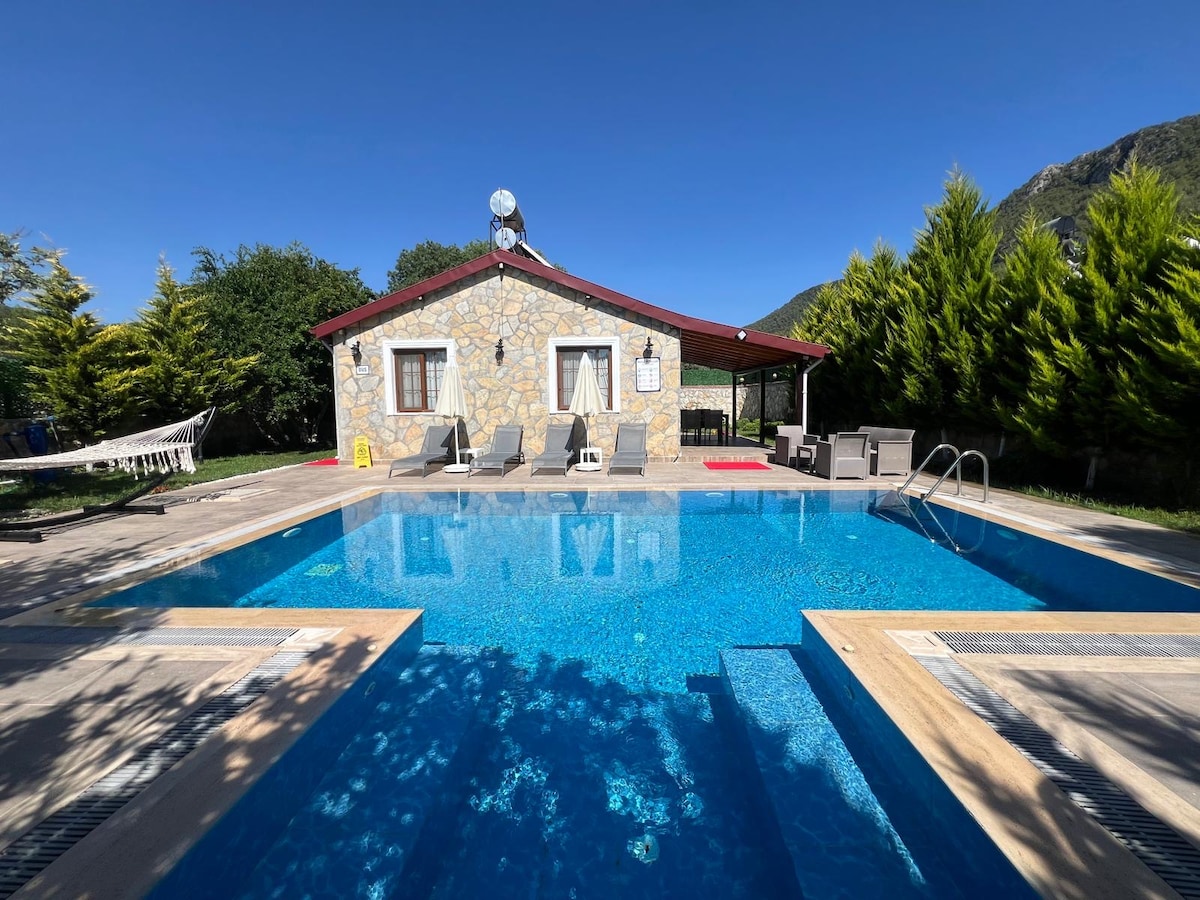 A stone villa is positioned beside a clear blue pool, with sun loungers arranged on a deck. A covered patio provides seating, surrounded by greenery. The vibrant sky is visible overhead, and the structure features several windows that bring natural light into the space.
