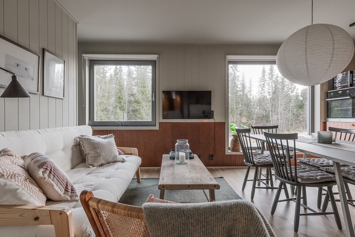 A bright living space features large windows allowing natural light and views of surrounding trees. A cozy sofa is paired with a wooden coffee table. A dining table with six chairs is positioned nearby, complemented by modern lighting and decorative elements.
