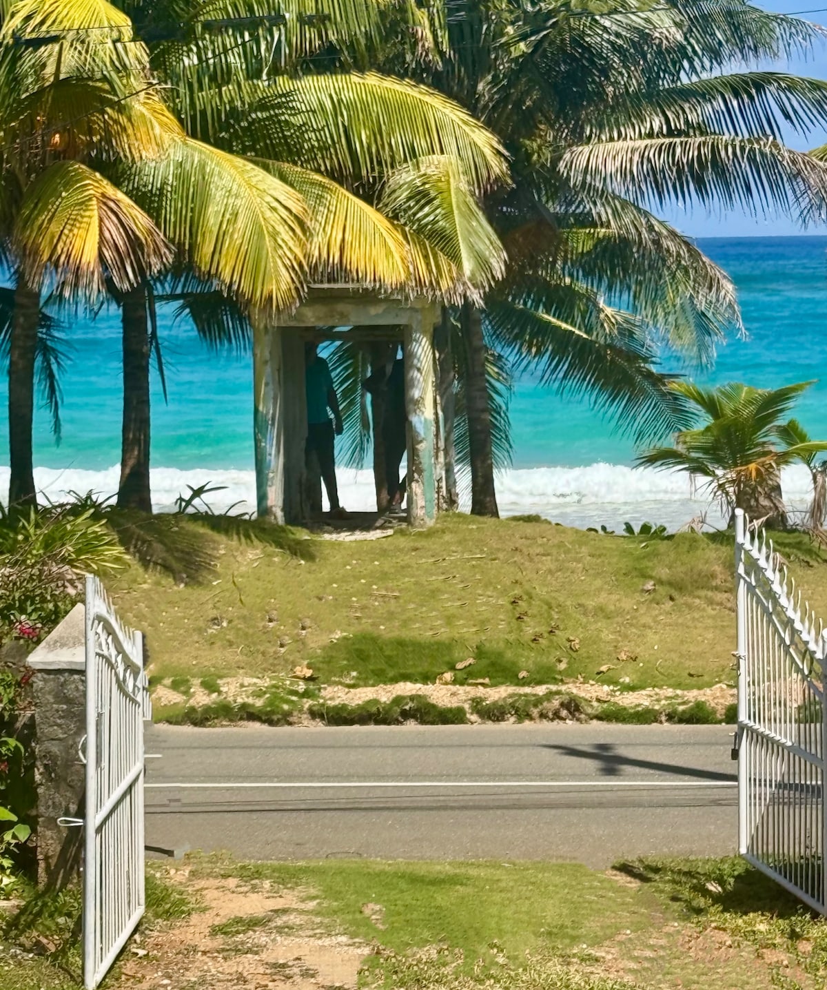 A pathway leads through a white gate towards a scenic view of the beach. Palm trees frame the opening, and turquoise waters can be seen beyond the grassy area. The vibrant greenery enhances the tropical setting.