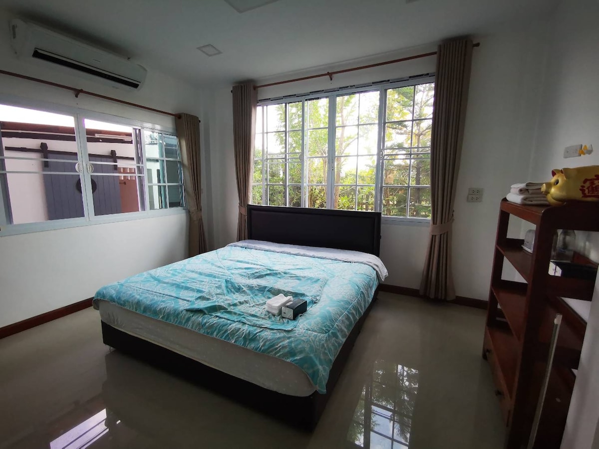 A spacious bedroom features large windows that allow natural light to fill the room. A comfortable bed with a light blue coverlet is centered against a dark headboard. A wooden shelf is positioned against one wall, enhancing the room's functionality.