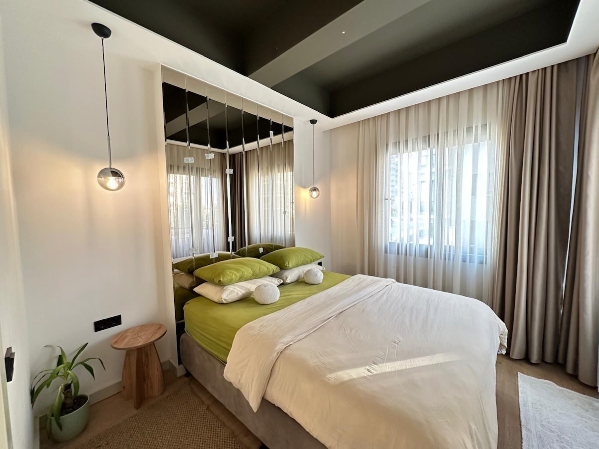 A cozy bedroom is featured with a spacious bed layered with green linens and multiple pillows. Soft natural light enters through sheer curtains, enhancing the serene ambiance. A small wooden table is positioned beside the bed, and a potted plant adds a touch of nature to the space.