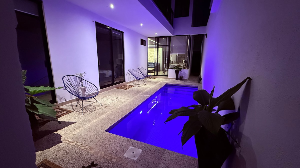 A private pool is featured within the serene indoor space, illuminated by soft purple lighting. Two modern lounge chairs are positioned beside the pool, complemented by leafy indoor plants that enhance the tranquil atmosphere.