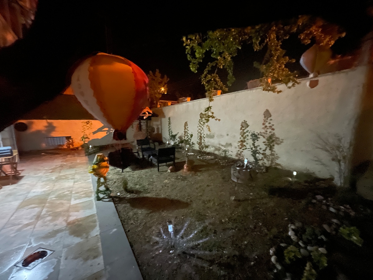 The garden features a spacious area with soft lighting and carefully arranged flower beds. A hot air balloon is visible overhead, adding a unique touch to the nighttime scene. Outdoor seating is present, creating a cozy spot for relaxation in the well-maintained space.