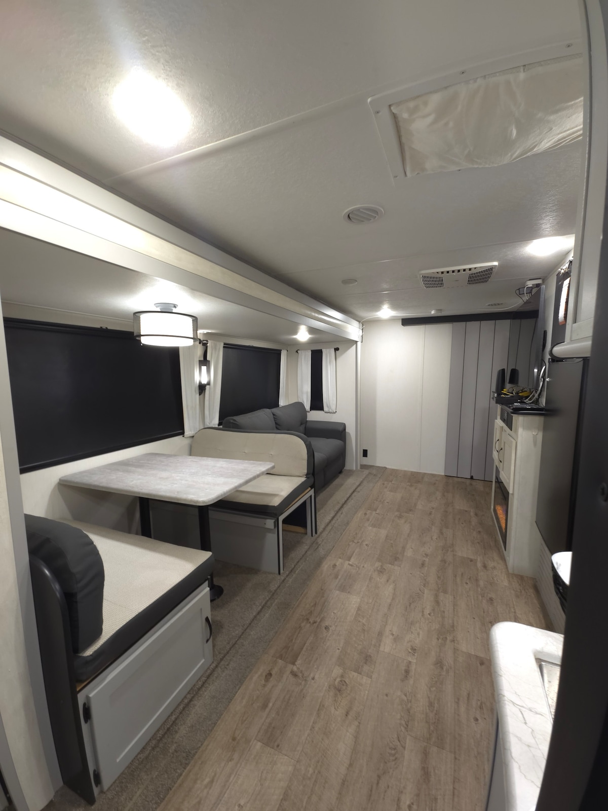 An open living area features two seating spaces with a table between them. Soft gray tones dominate the furnishings. Windows, covered with dark blinds, allow for controlled lighting. A kitchenette area with modern appliances is situated in the background, completing the space.