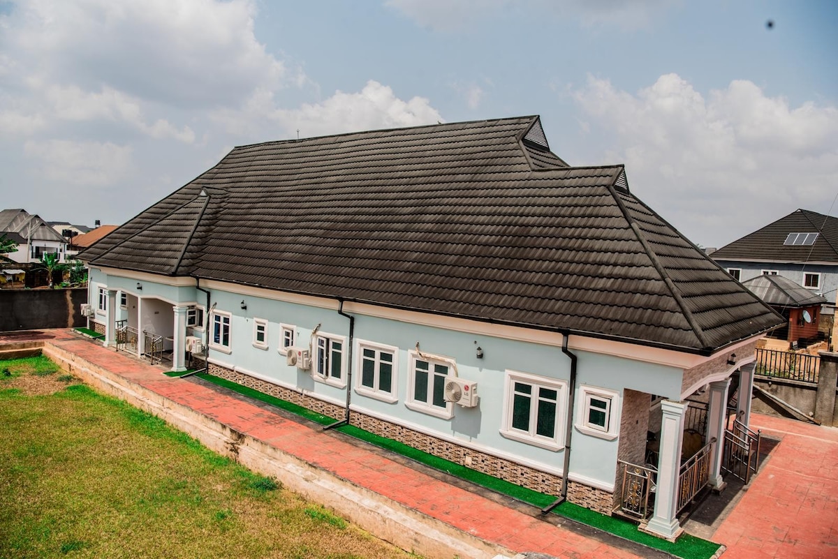 The exterior of a spacious two-story apartment is displayed, featuring a gabled roof and multiple windows. Air conditioning units are noticeable on the walls, providing comfort. The surrounding green lawn extends to a paved outdoor area, with a clear blue sky above, contributing to the overall openness.