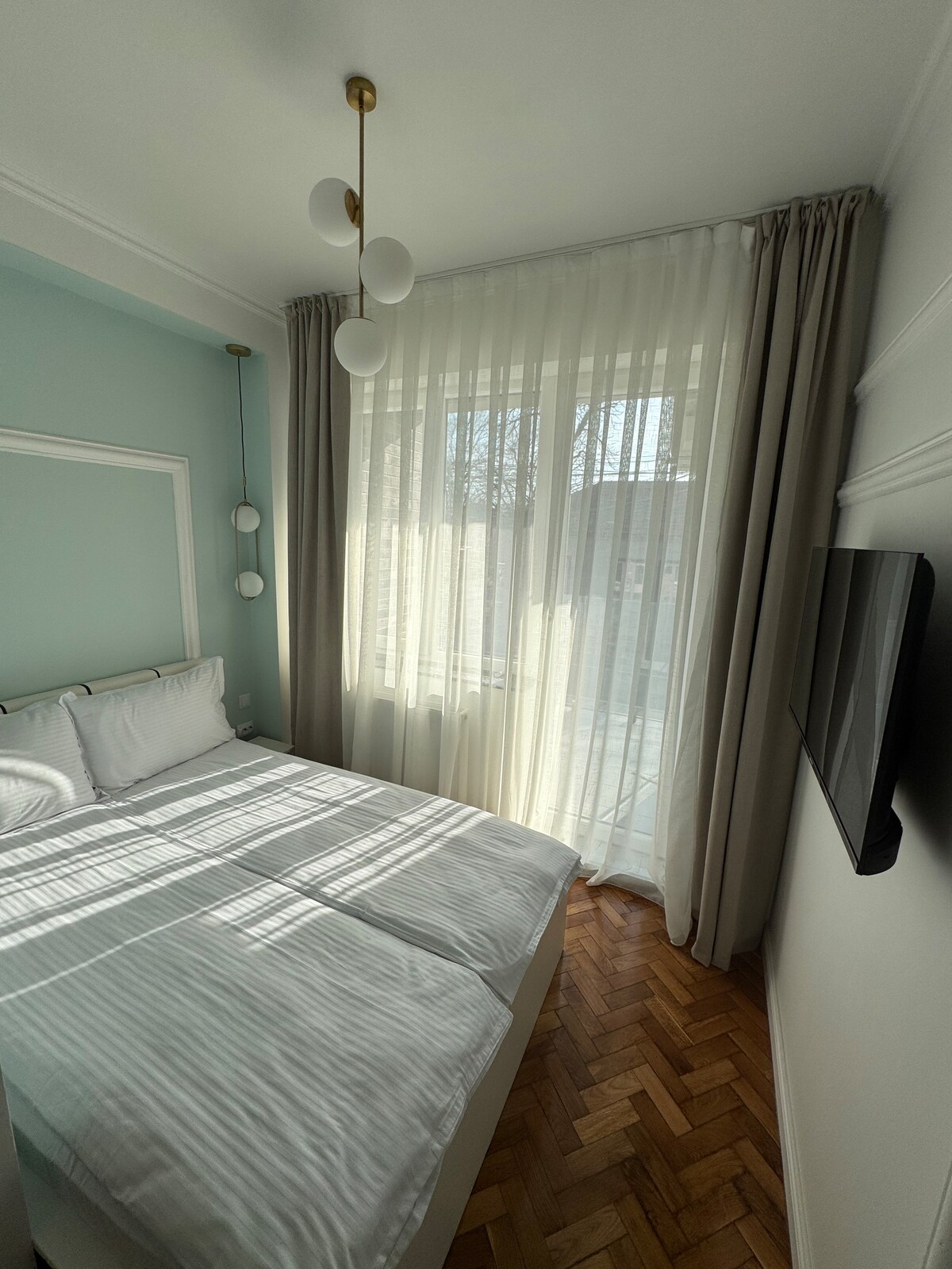 A bright bedroom features a spacious bed with light bedding, positioned near a large window. Soft curtains diffuse natural light, creating a serene environment. A television is mounted on the wall opposite the bed, and wooden flooring adds warmth to the space.