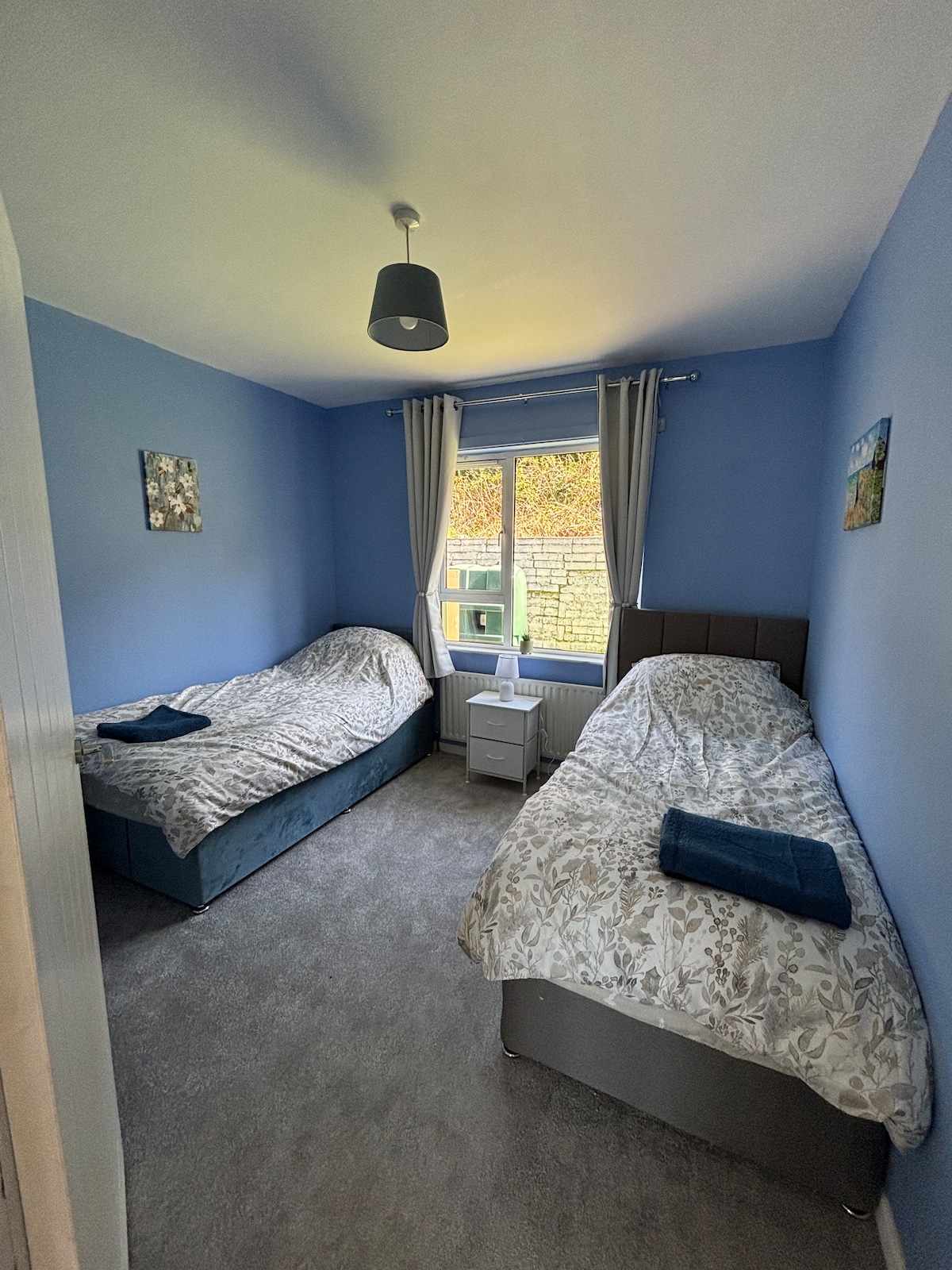 A twin bedroom featuring two separate beds with floral-patterned bedding. Soft grey carpet covers the floor. A small bedside table sits between the beds, supporting a lamp. Light enters through a window framed by light-colored curtains, creating a calm atmosphere in the room.