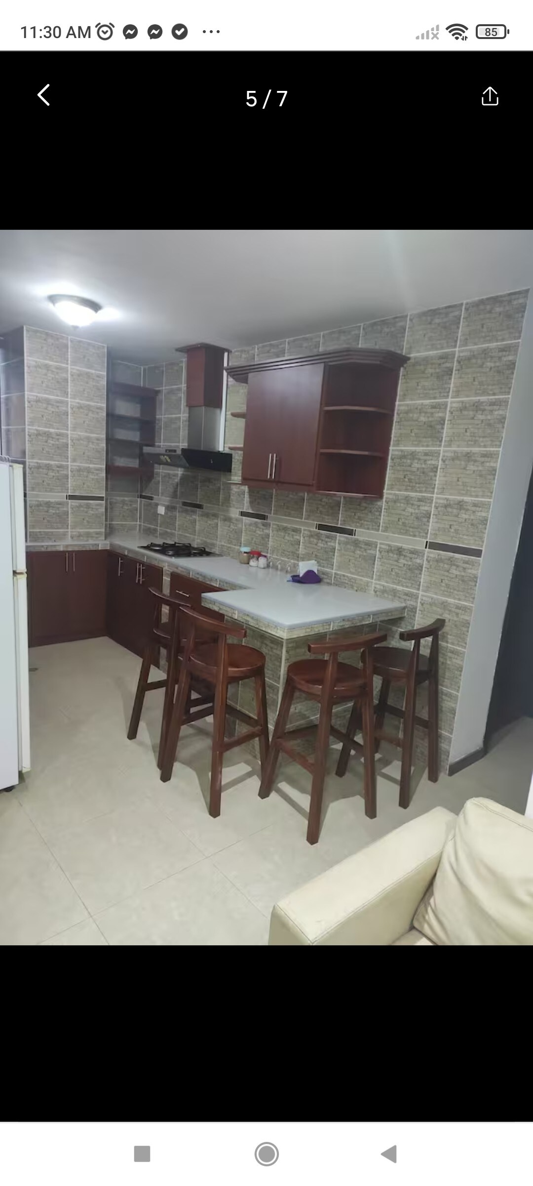 A modern kitchen area features dark wood cabinetry and a granite countertop. A small dining table with four wooden stools is positioned near the center. The walls are adorned with stone tiles, contributing texture to the space, while a refrigerator stands against one side.