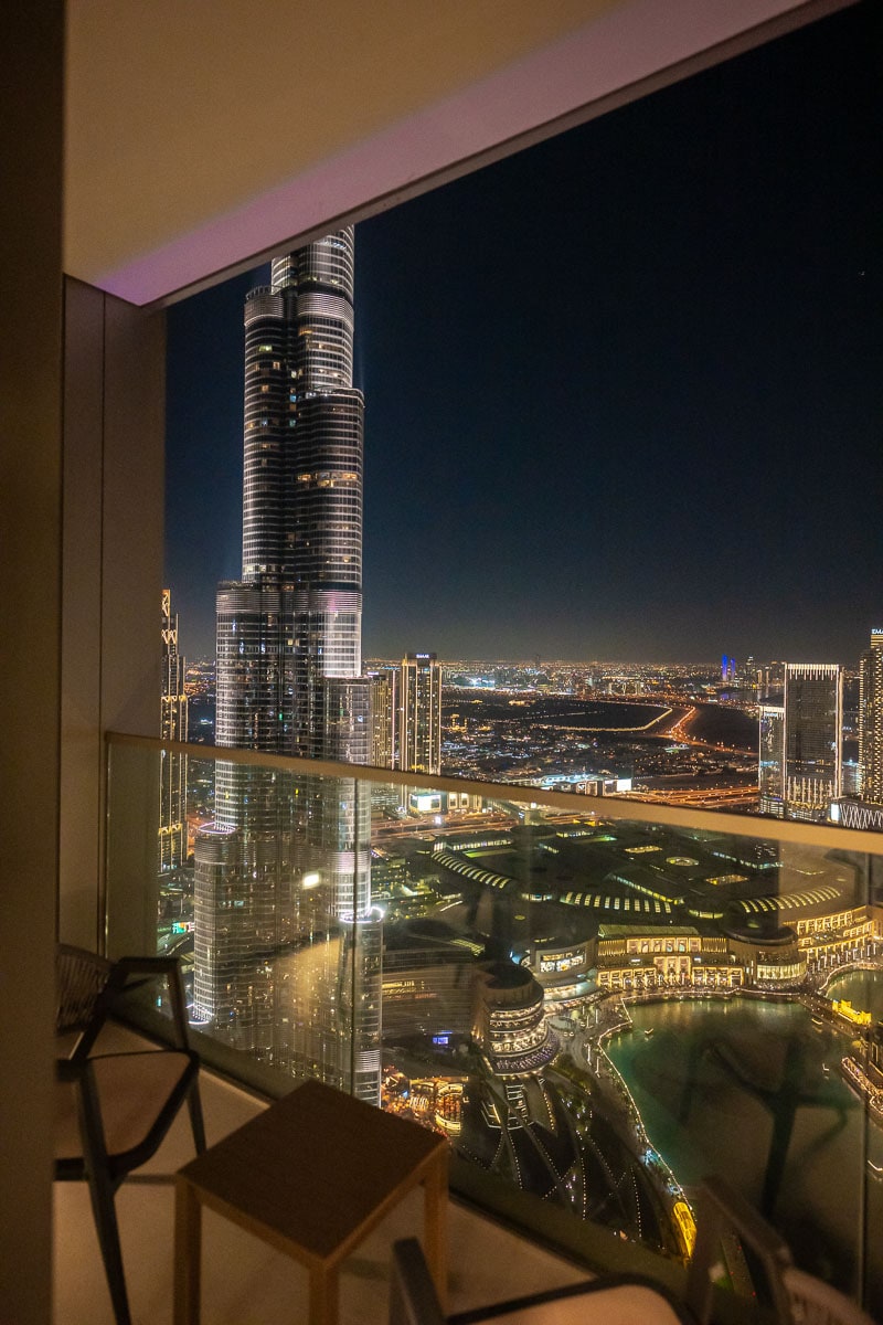 A nighttime view from the balcony highlights the impressive silhouette of the Burj Khalifa, illuminated against a dark sky. City lights sparkle below, showcasing the vibrant downtown area, with the Dubai Fountain and surrounding architecture visible in the foreground.