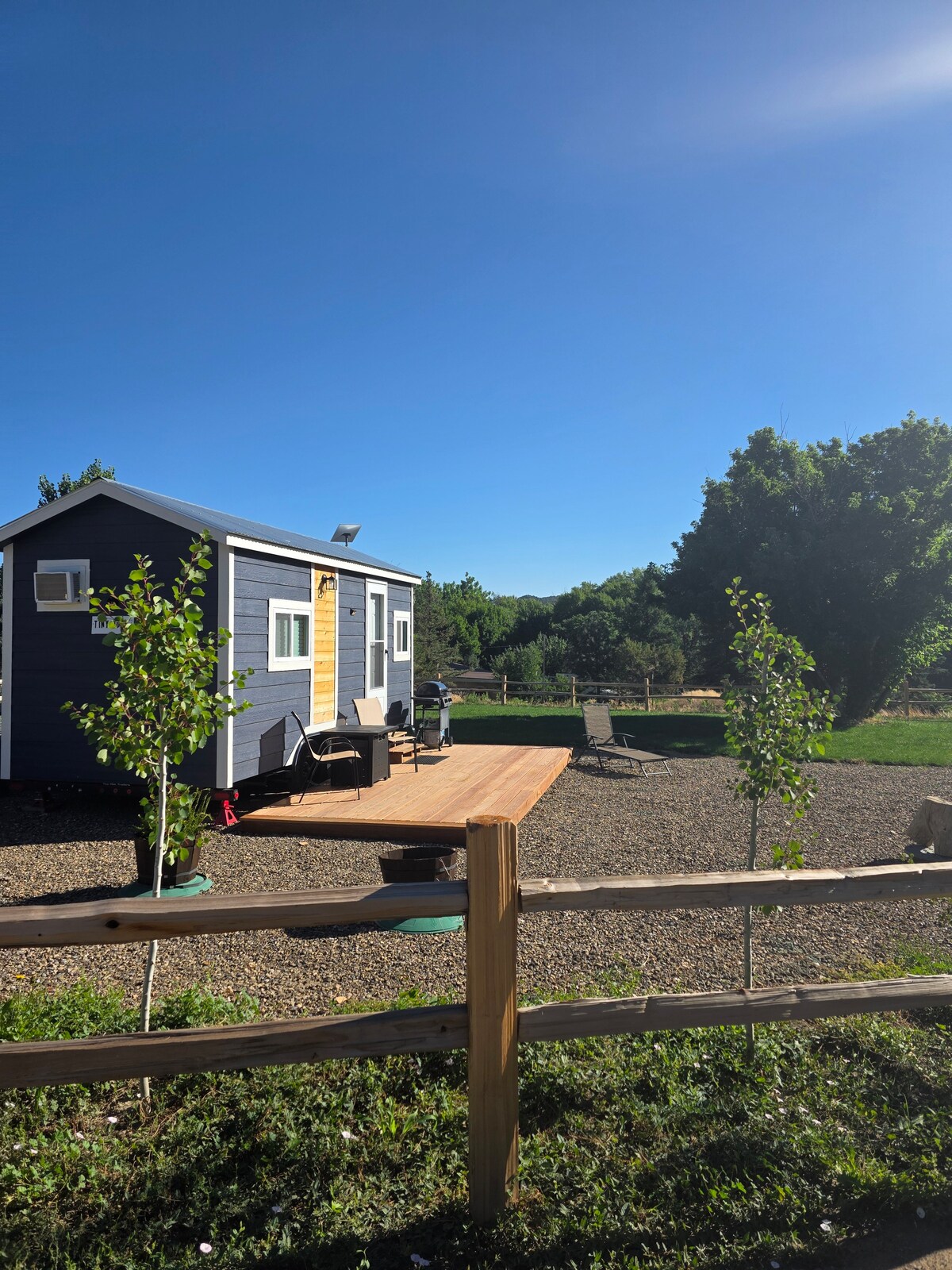 A tiny home is set against a clear blue sky, featuring a wooden deck adorned with seating. Lush greenery surrounds the property, while a fenced area provides privacy. A gravel pathway leads to a landscaped yard, enhancing the serene atmosphere of the location.