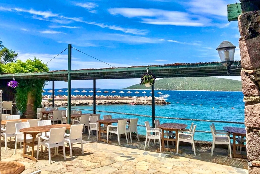 A seaside dining area features light wooden tables and white chairs set on a stone patio. The view includes vibrant blue waters and clear skies, with umbrellas lining the shore in the distance. Lush greenery is seen along the edges, enhancing the tranquil setting.