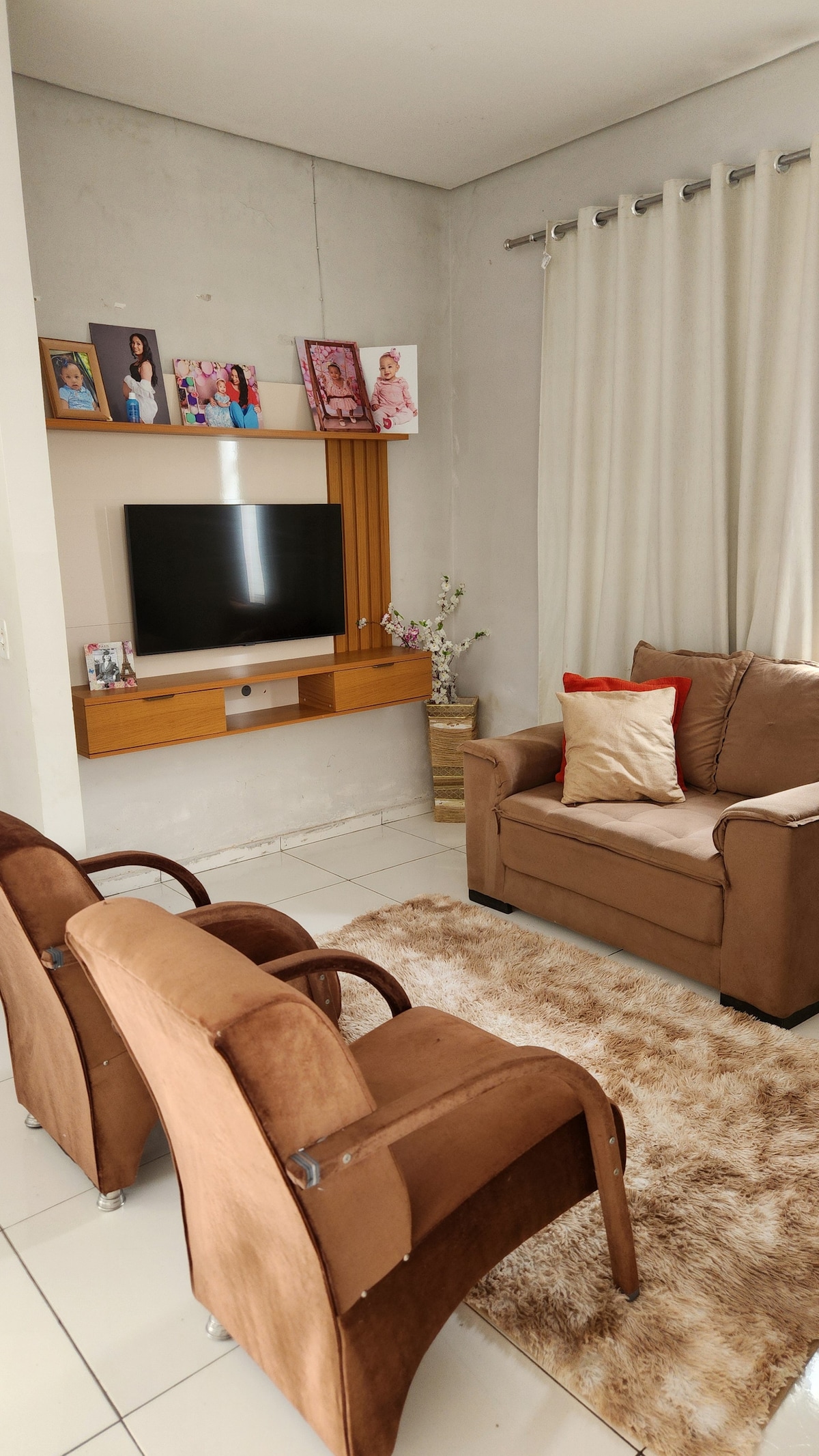 A cozy living area features two armchairs positioned across from a brown sofa, complemented by decorative pillows. A medium-sized television is mounted on a wooden wall unit, flanked by family photographs. Soft curtains frame a window, and a light-colored area rug adds warmth to the space.