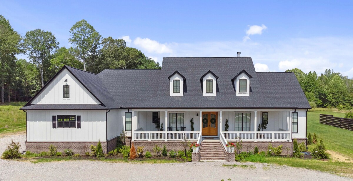 The exterior of a modern farmhouse is showcased, featuring a symmetrical design with multiple gabled roofs. A welcoming entrance is framed by a broad porch adorned with potted plants, complemented by expansive windows that invite natural light. Lush greenery surrounds the property, enhancing its natural setting.