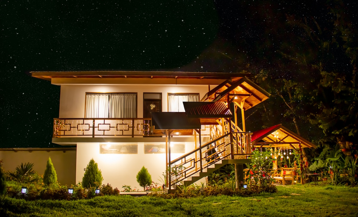 A two-story private residence is illuminated softly against a starry night sky. The structure features wooden accents and a welcoming porch with railings. Surrounding greenery and subtle garden lights enhance the tranquil outdoor setting.