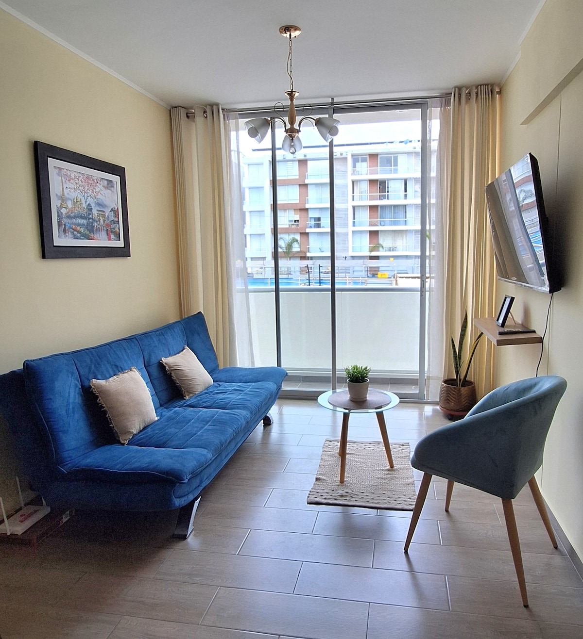 A bright living area features a blue sofa positioned near a large glass window, allowing natural light to fill the space. A small coffee table is positioned in front of the sofa, with a potted plant nearby. A television is mounted on the wall across from the seating.