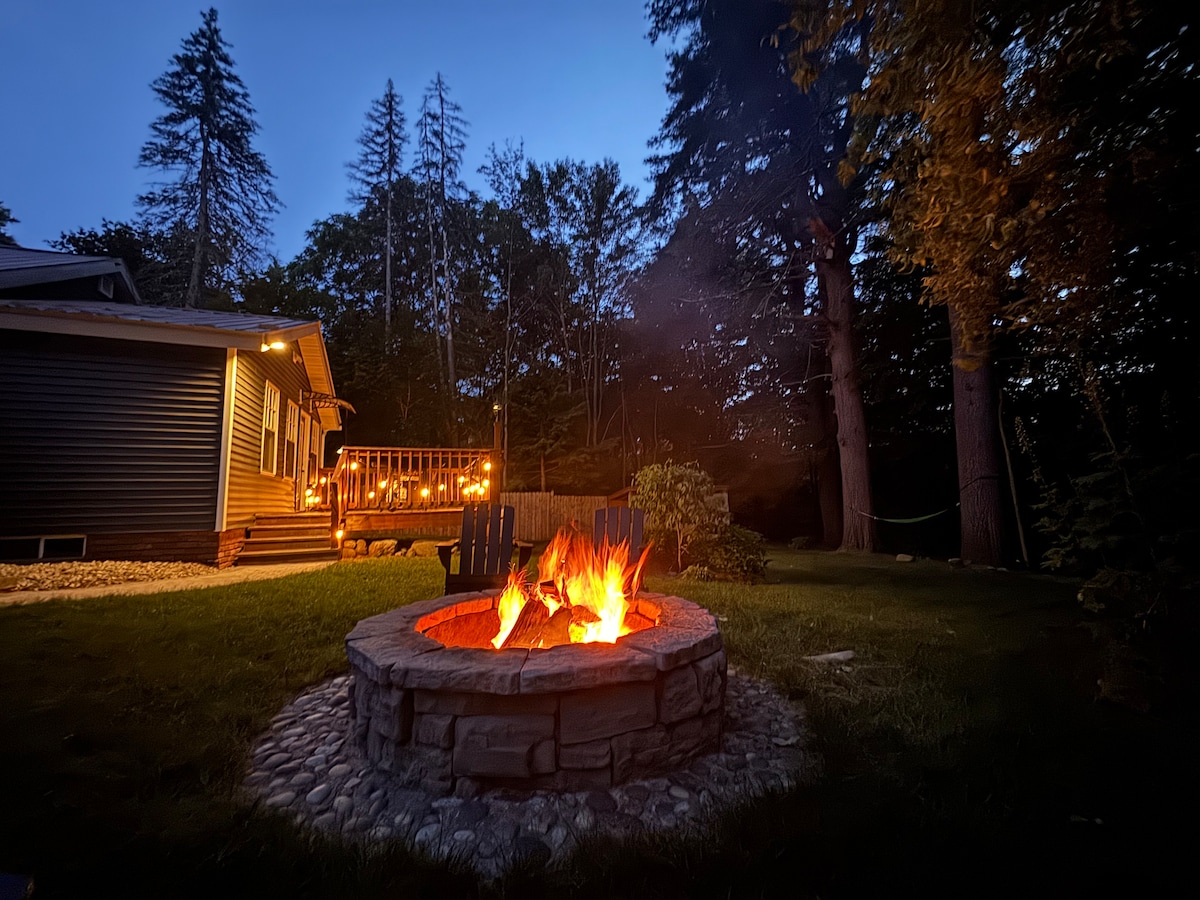 A cozy fire pit is surrounded by stonework and adirondack chairs, glowing warmly in the evening. Soft lighting from the nearby deck creates a welcoming atmosphere against the backdrop of thick trees and a quiet home.