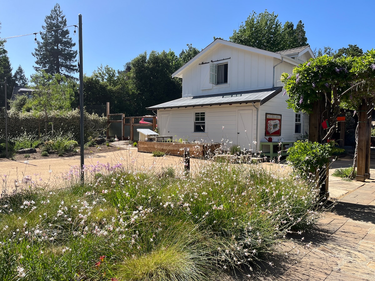 A white two-story studio is set amidst a garden featuring native flora and tall trees. Sunlight illuminates the outdoor area, highlighting the natural surroundings. A gravel pathway leads to the private entry, while a wooden structure provides additional space for leisure activities.