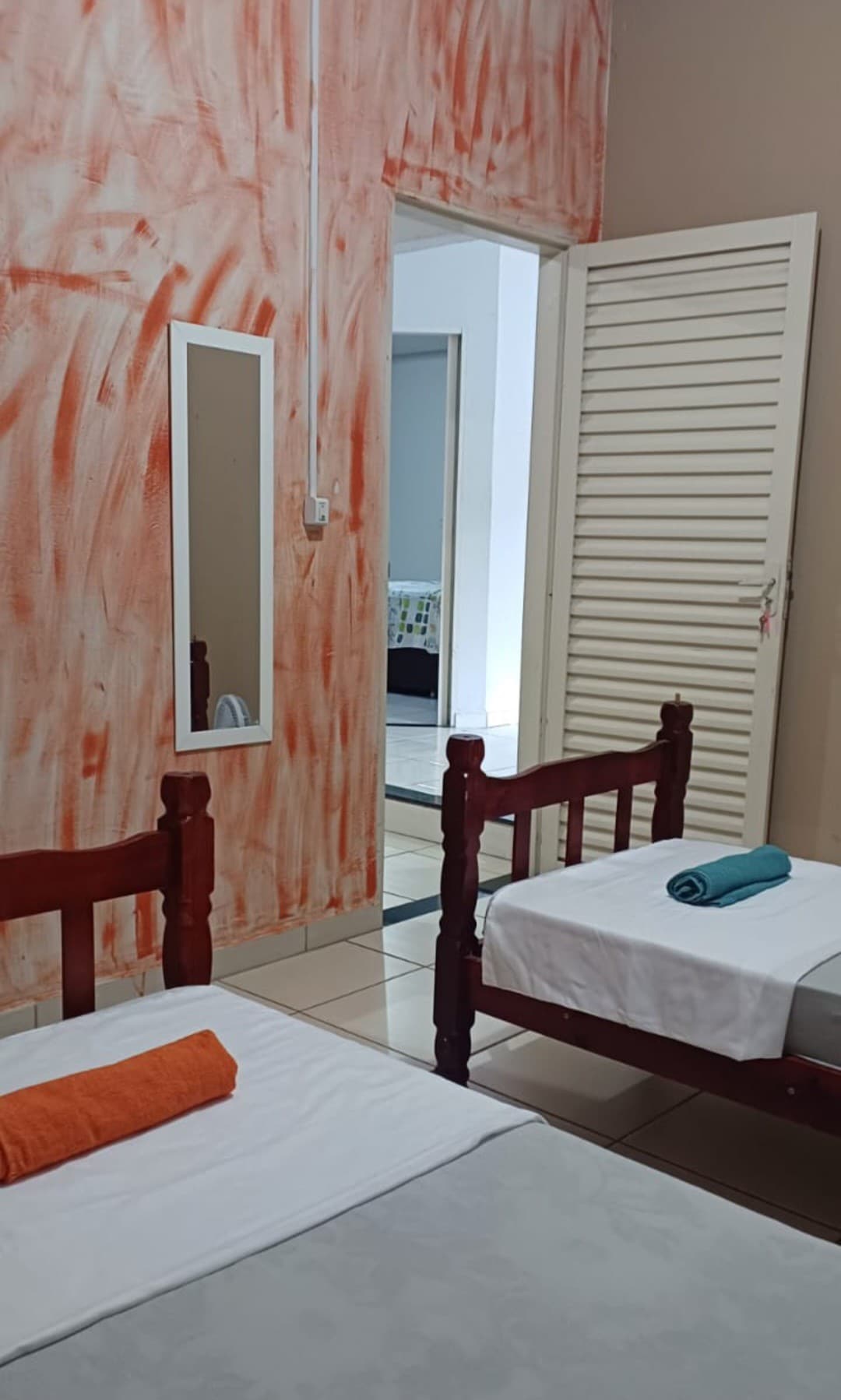 A simple room showcases two wooden beds, each featuring fresh linens. An orange accent towel is placed on one bed, while a teal towel is neatly rolled on the other. A floor mirror and a partially open white door reveal additional space beyond.