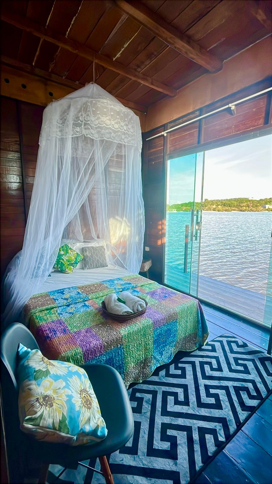A cozy sleeping area features a colorful quilt draping over the spacious bed, complemented by decorative pillows. Natural light streams in through the glass door, which opens to a water view. A small chair with floral accents is positioned beside the bed, adding to the tranquil setting.