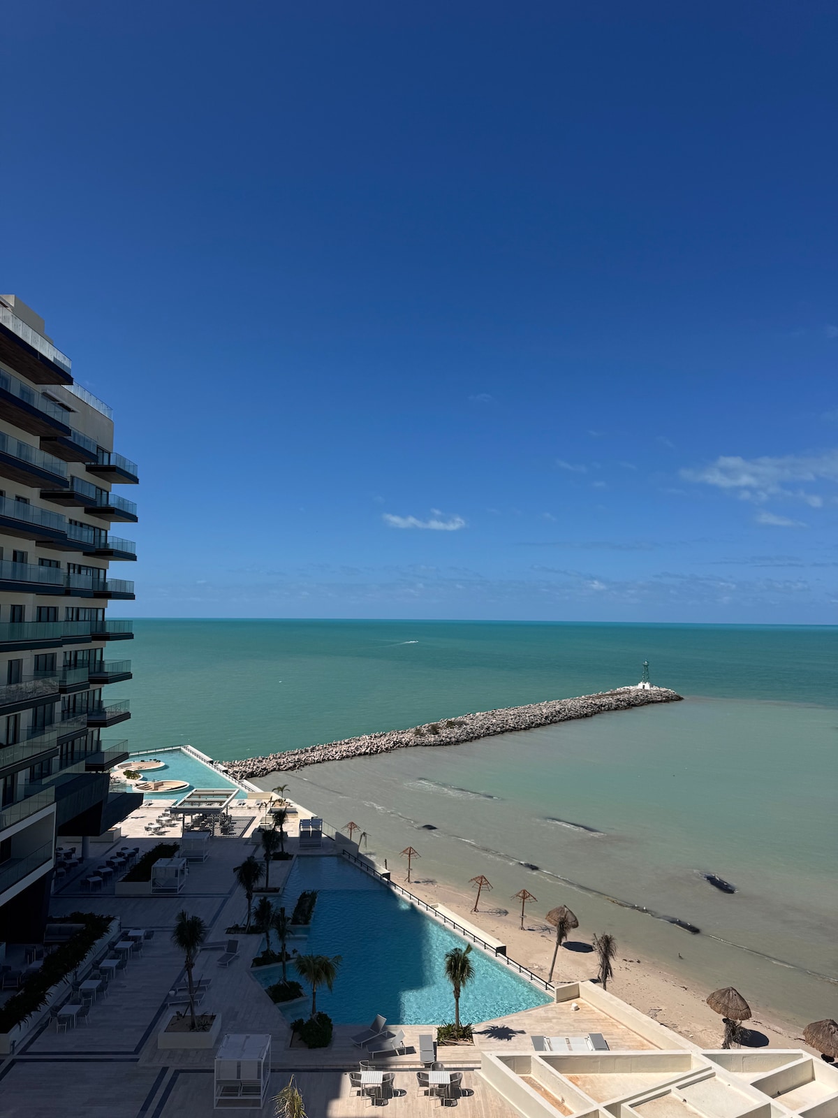A panoramic view captures the serene beach and calming ocean waters, with gentle waves lapping at the shore. A sleek pool area is visible in the foreground, complemented by a jetty extending into the sea. The sky is clear, enhancing the tranquil atmosphere.
