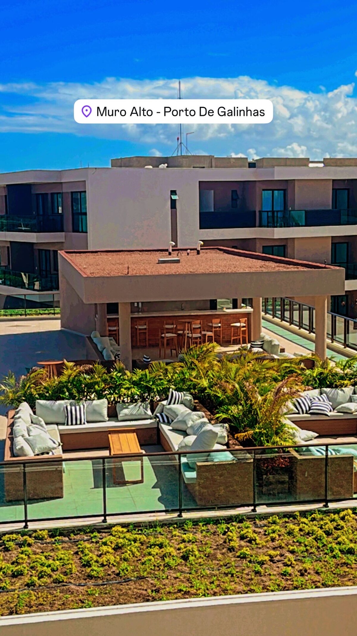 A spacious outdoor lounge area is surrounded by greenery, featuring comfortable seating arrangements with light-colored cushions. A sheltered dining space is visible nearby, set against a backdrop of modern architecture. The bright blue sky enhances the inviting atmosphere of the communal space.