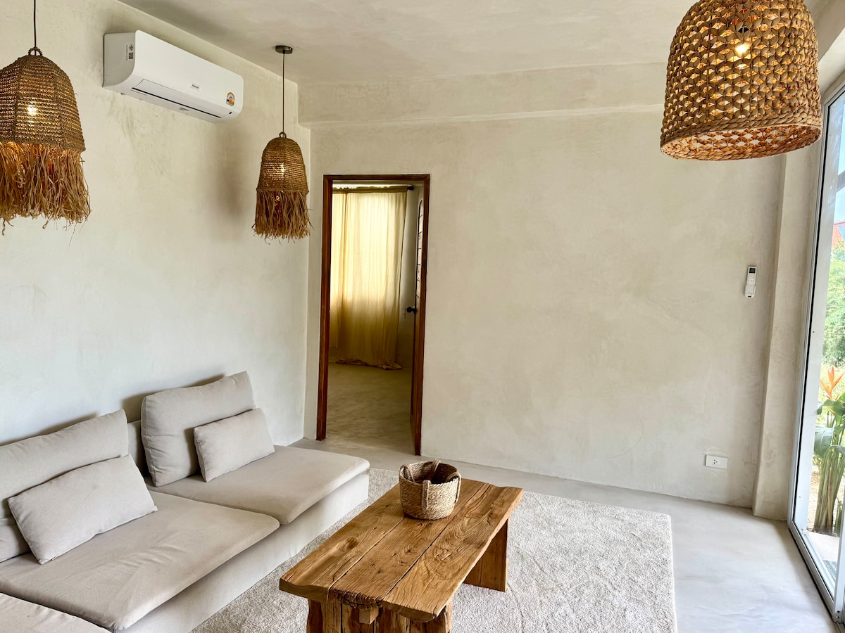A natural light-filled living area is furnished with a soft, neutral-toned sofa and a rustic wooden table. The walls exhibit a textured finish, while woven pendant lights create a calming ambiance. A doorway leads to another room, enhancing the spacious feel.