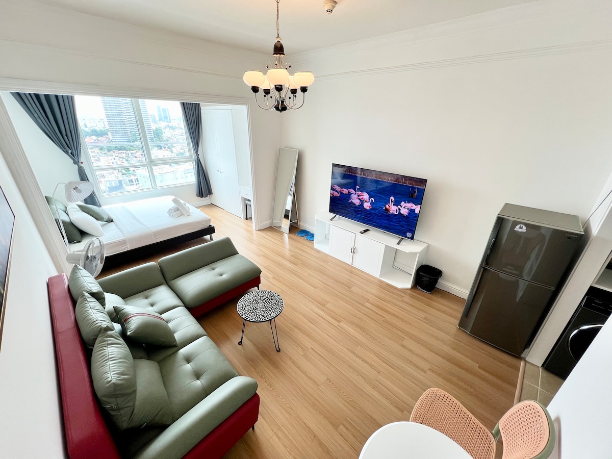 An open living space is showcased, featuring a green sectional sofa and a black coffee table. A large TV is mounted on the wall opposite a spacious bed. Natural light streams through large windows, enhancing the inviting atmosphere. A dining area with two chairs is also visible.