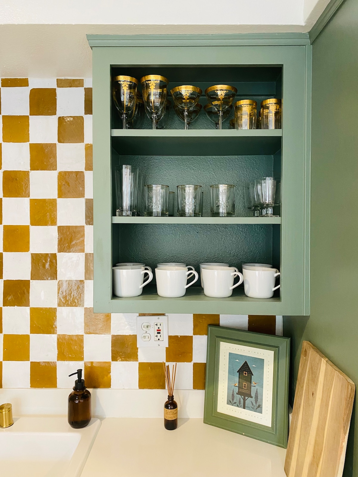 A green cabinet displays an array of glassware, including ornate gold-accented glasses and clear tumblers. White mugs are stacked neatly on the lower shelf. A framed art piece and a wooden cutting board are placed on the countertop beneath the cabinet, enhancing the kitchen's design.