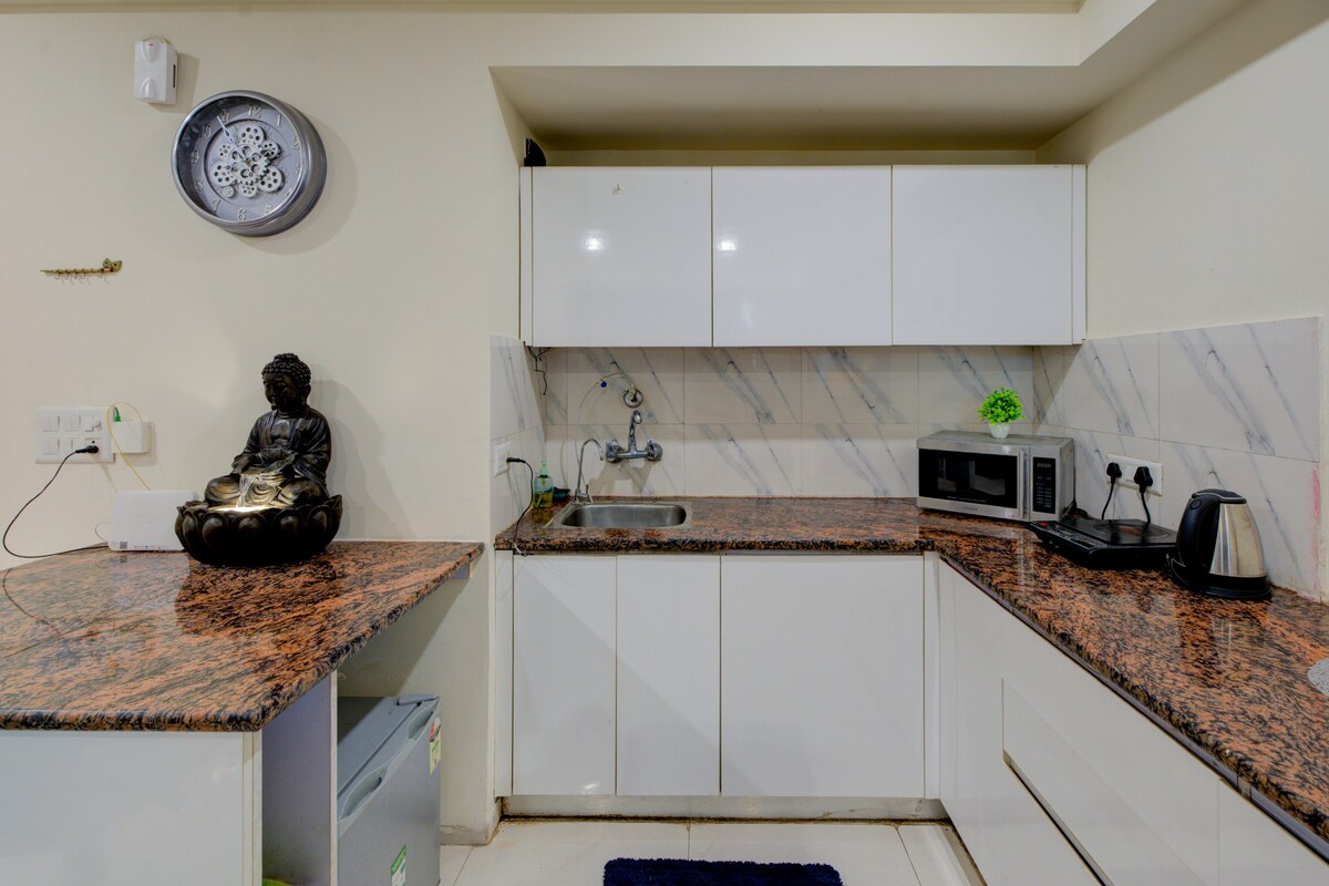 A modern kitchen features sleek white cabinetry and a polished granite countertop. Essential appliances, including a microwave and kettle, are present. Decorative elements, such as a statue and wall clock, add character. The space is designed for functionality and convenience.