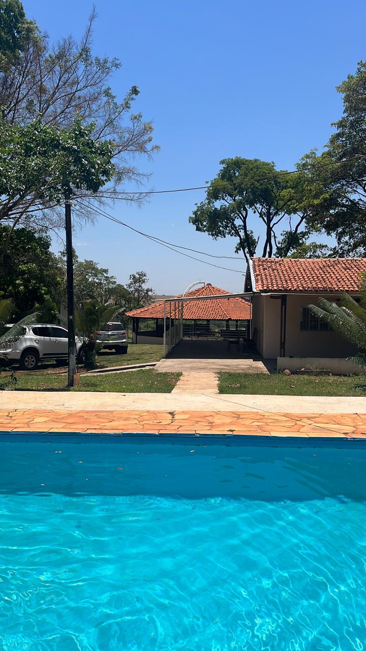 The image showcases a clear blue swimming pool in the foreground, with a view of a covered outdoor area featuring a pergola in the background. Lush greenery surrounds the space, and a pathway leads to a single-story house with a tiled roof.