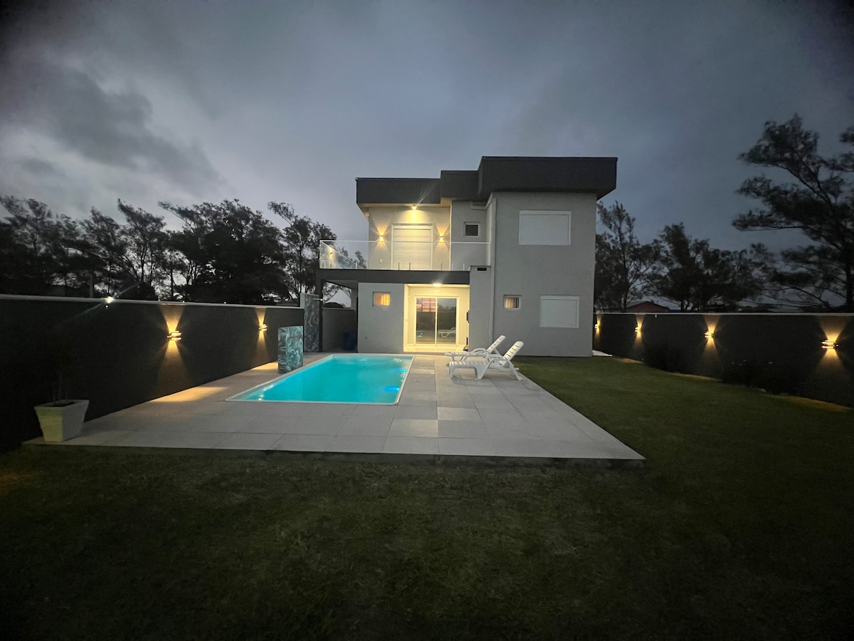 A modern two-story house is illuminated under a twilight sky, featuring a sleek outdoor pool. Lounge chairs are positioned by the poolside on a tiled patio, surrounded by a well-maintained grassy area. Soft lighting highlights the exterior walls, creating a serene atmosphere.