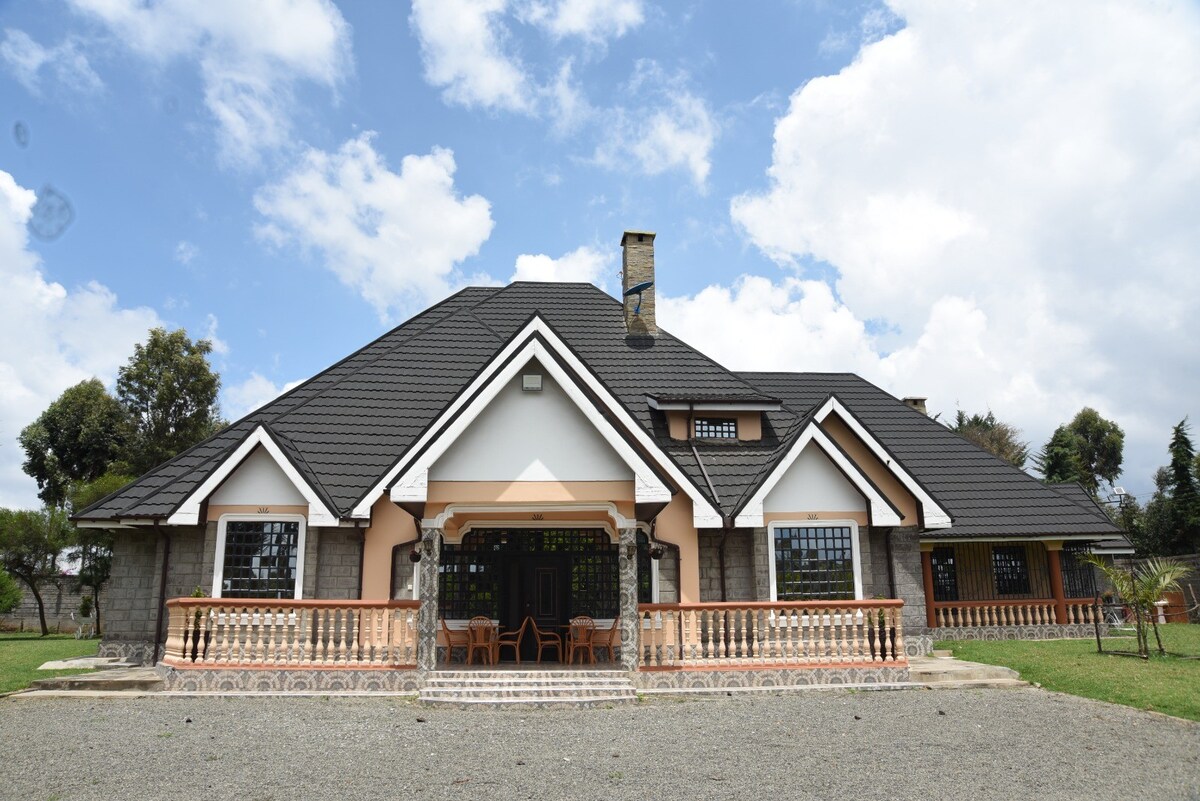 The two-story home features a combination of stone and white exterior walls, topped with a dark sloped roof. A covered porch is framed by four wooden chairs, providing a view of the surrounding green lawn and trees under a partly cloudy sky.