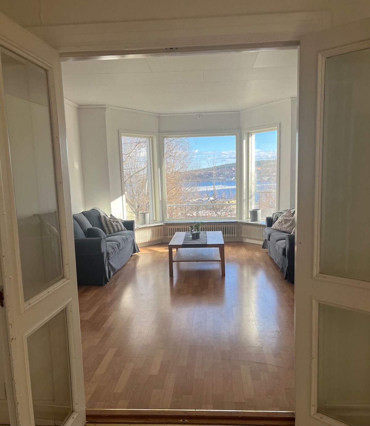 An inviting living room is viewed through glass-paneled doors. It features large windows that offer panoramic views of the landscape. Two sofas are positioned around a central coffee table, with natural light flooding the space from the windows, enhancing the overall brightness.