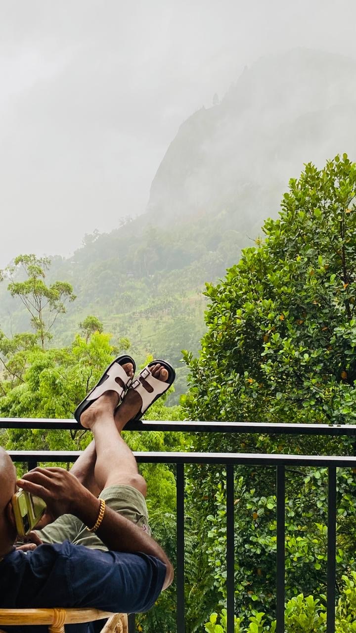 A person relaxes on a balcony, with legs propped up, showcasing comfortable footwear. Lush greenery surrounds the area, leading towards a mountainous landscape partially shrouded in mist. The serene environment reflects a peaceful natural setting, inviting moments of tranquility.